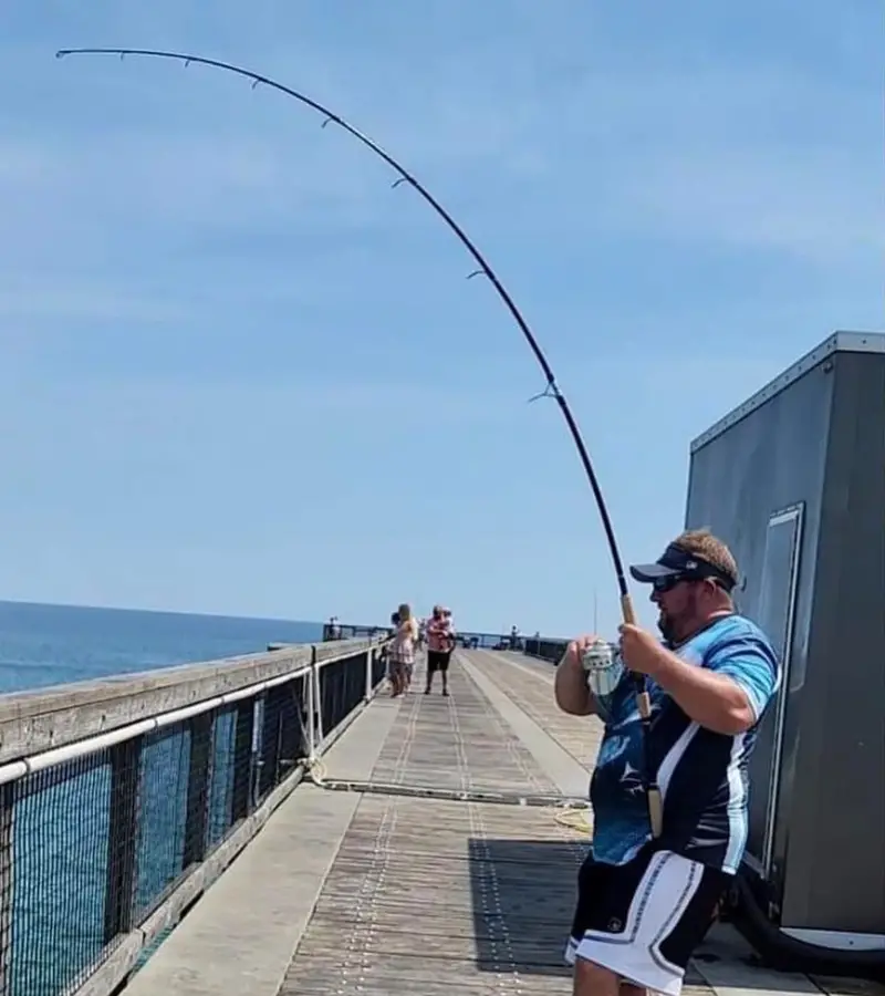 Photo of an angler with a long fishing rod on a pier fighting a big fish